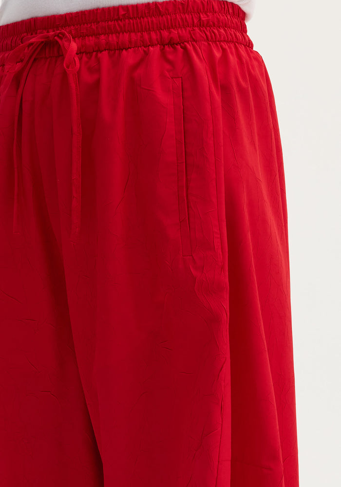 Balloon track pants in red, featuring an elasticated waistband with drawstring, slit side pocket, and crinkled texture for a voluminous, stylish look.