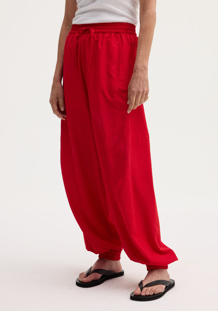 Balloon track pants in red with elasticated waistband and cuffs, featuring front welt pockets and a voluminous leg, styled with black flip-flops.