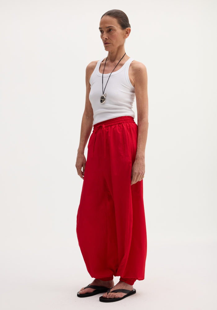 Balloon track pants in red with voluminous legs, elasticated waistband, and hems. Features front welt pockets. Model wears pants with a white tank top and flip-flops.