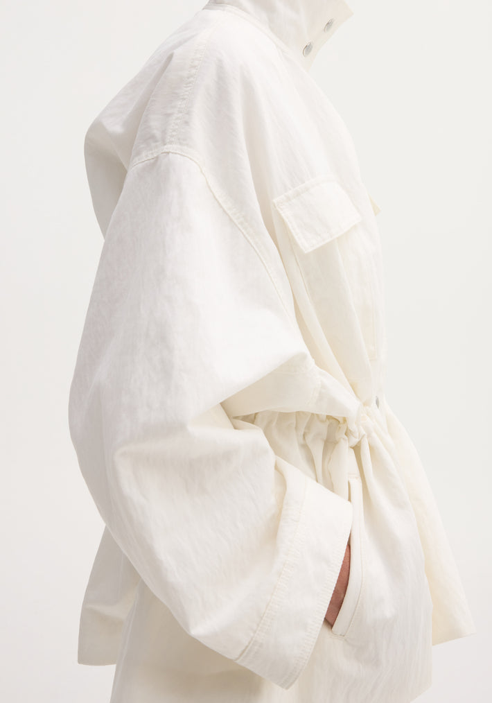 Sculptural safari jacket with oversized sleeves, snap-button stand collar, flap chest pocket, and drawstring waist, showcasing a utilitarian yet clean silhouette.