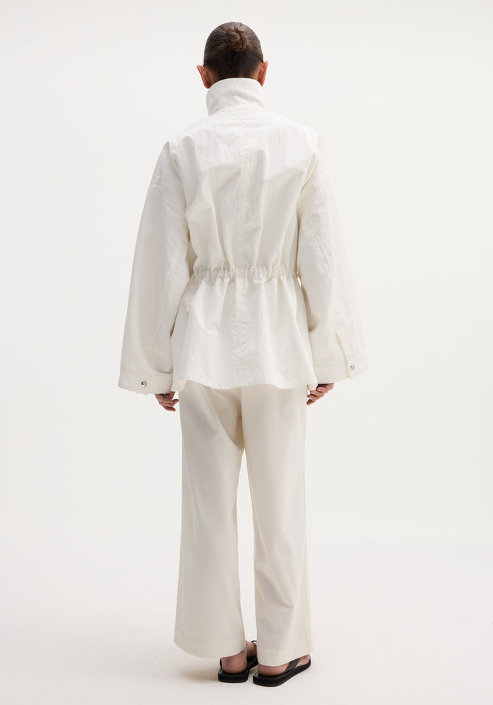 Back view of a person in a Sculptural safari jacket featuring a high collar, oversized pockets, and elasticated waist, paired with matching trousers and black sandals.