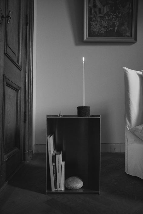 Round Candle Holder on minimal side table, crafted from lava stone, featuring a tall candle. Books and a stone accent the scene's serene, sparse design.