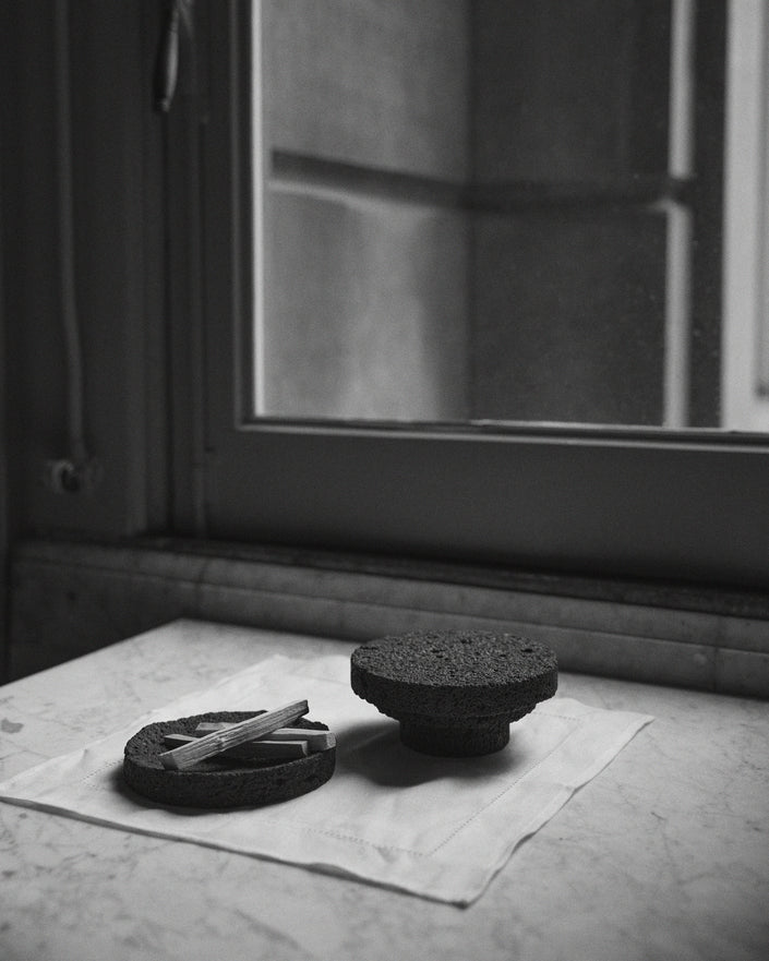 Round Tray made of textured lava stone, one on a pedestal, another holding wooden sticks, showcasing its handcrafted uniqueness on a white cloth.