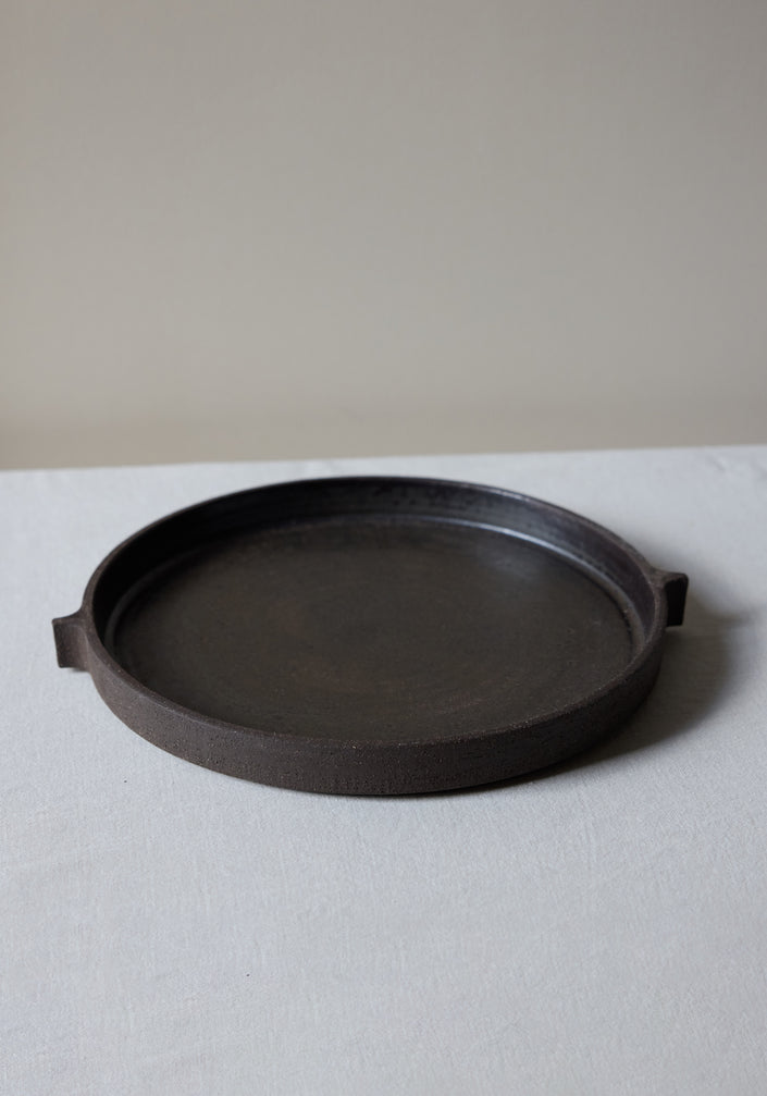 Ceramic Serving Plate | soil: A wide, flat, low-walled circular plate with a rough finish, reminiscent of brutalist design, rests on a light cloth surface.