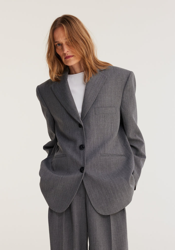 Person wearing an oversized blazer in grey melange from the Frame collection, featuring broad shoulders and paired with matching wide-leg trousers, hands in pockets.