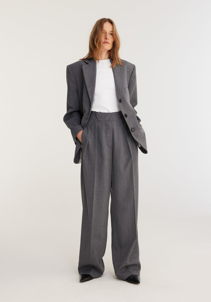 Model wearing Oversized blazer | grey melange with broad shoulders, high-waisted wide-leg trousers, and a white T-shirt, showcasing a minimal, androgynous power suit.