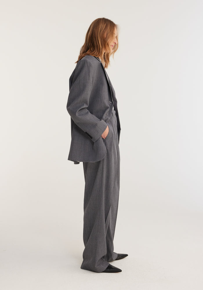 Person wearing an oversized grey melange blazer from Italian mill Bisentino, paired with matching wide-leg trousers and pointed black shoes, standing in profile.