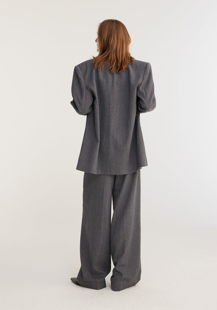 Person in oversized grey melange blazer, featuring padded shoulders, paired with matching wide-leg trousers, standing in a studio. Part of Frame's permanent collection.