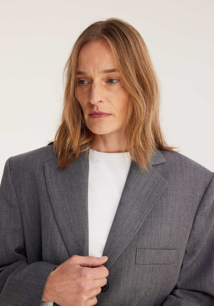 Woman wearing an oversized blazer in grey melange, tailored from a wool-blend fabric by Italian mill Bisentino, holding the lapel with a clean, minimalist background.
