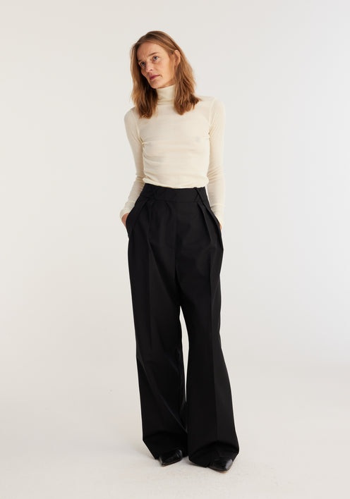 Woman wearing Fine merino turtleneck | off-white, paired with high-waisted black trousers, hands in pockets, showcasing a classic, fitted silhouette ideal for layering.