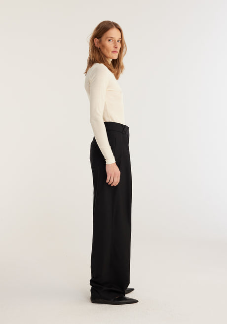 Woman wearing Fine merino turtleneck | off-white, paired with high-waisted black trousers, hands in pockets, showcasing a classic, fitted silhouette ideal for layering.