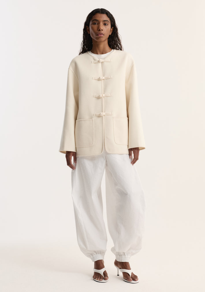 Model wears Double-faced wool pankou jacket with frog-toggle closures, embroidered edges, and patch pockets, paired with loose crinkled pants and heeled sandals.
