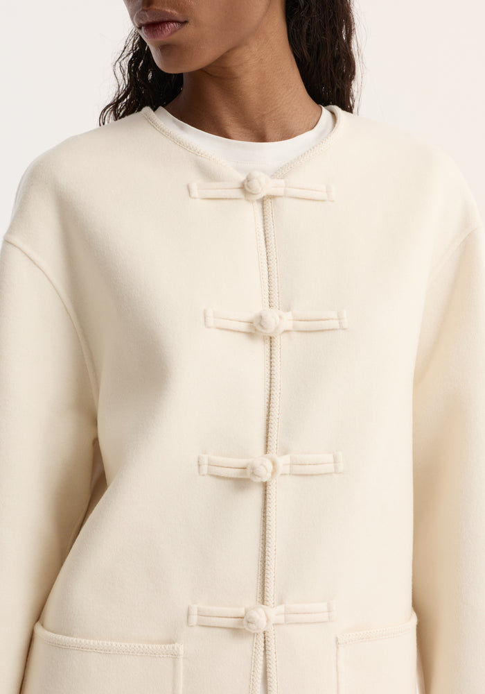Double-faced wool pankou jacket in off white, featuring pankou knot closures, embroidered edges, and patch pockets, worn over a white T-shirt.