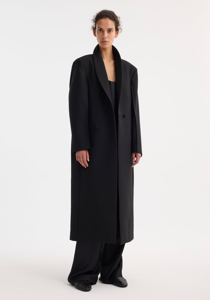Person wearing the Signature opera coat with a tailored black overcoat, single-button closure, softly rolled collar, and wide-leg trousers, exuding a minimalist, elegant style.