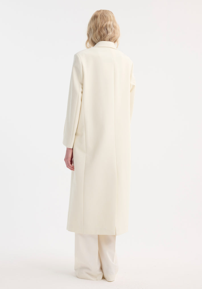 Signature opera coat | ivory with a structured silhouette, double-breasted lapels, and back slit, worn by a model with wavy blond hair, showcasing timeless elegance.