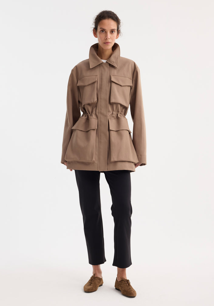 Model wearing the Field jacket with waist adjuster | clay, showcasing a structured collar, oversized bellows pockets, and adjustable waist, paired with black trousers and brown shoes.