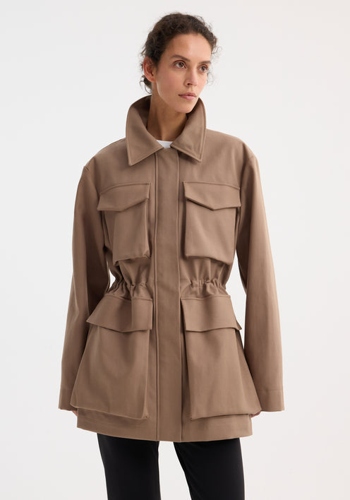 Model wearing the Field jacket with waist adjuster | clay, showcasing a structured collar, oversized bellows pockets, and adjustable waist, paired with black trousers and brown shoes.