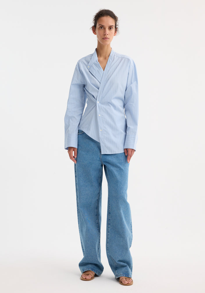 Model wearing the Overlap striped shirt in fine blue stripe with an asymmetrical front, paired with straight mid-blue jeans and tan slide sandals.