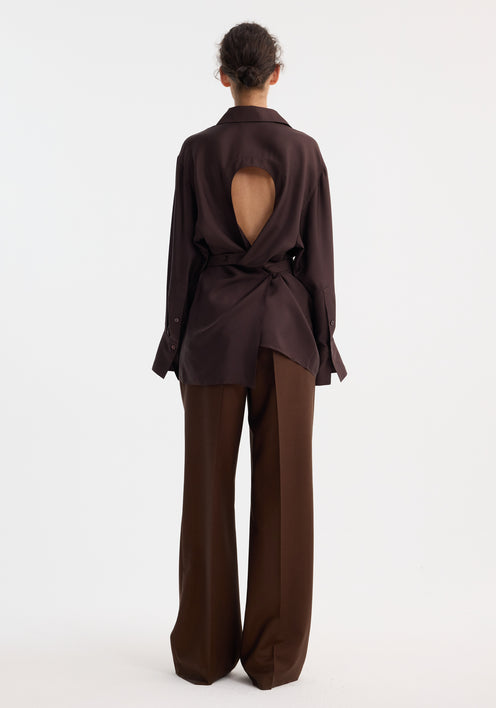Woman wearing a deconstructed silk shirt in mahogany, featuring an open back and versatile tie design, paired with matching trousers and beige pointed-toe shoes.