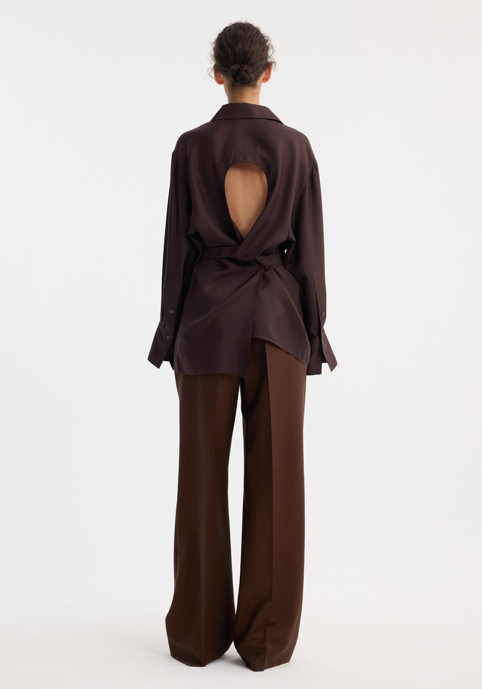 Woman wearing a deconstructed silk shirt in mahogany, featuring an open back and versatile tie design, paired with matching trousers and beige pointed-toe shoes.