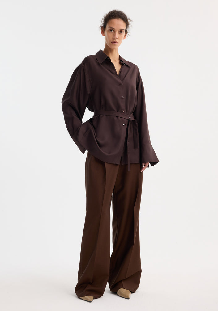 Woman wearing a deconstructed silk shirt in mahogany, featuring an open back and versatile tie design, paired with matching trousers and beige pointed-toe shoes.