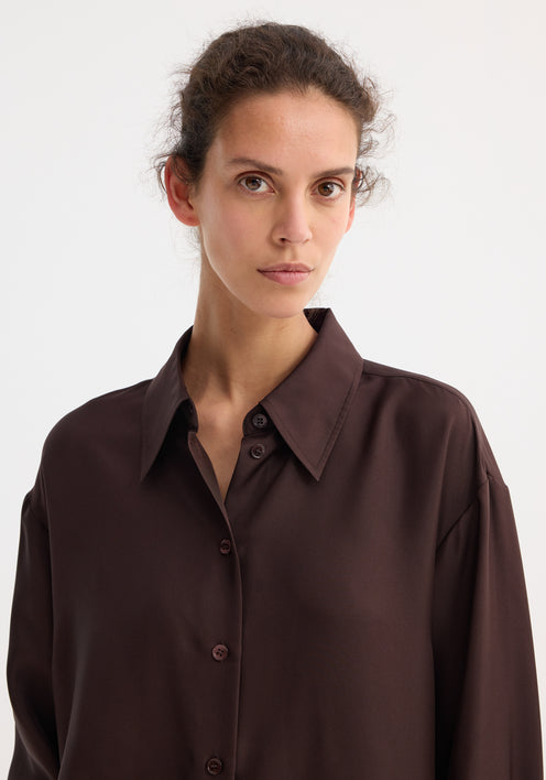 Woman models the Signature elongated silk shirt in mahogany, oversized with a statement collar, paired with matching wide-leg trousers and tan slide sandals.