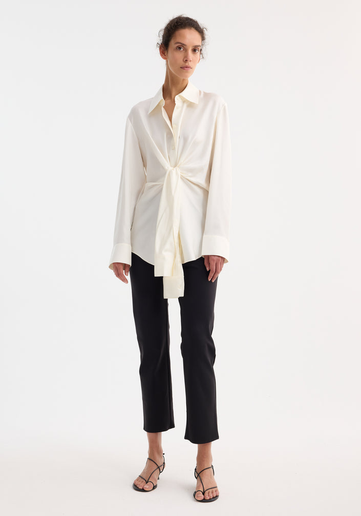 Model wearing Silk knotted shirt | cream, featuring a twisted front design, paired with slim black pants and black strappy heels, showcasing an elegant, refined look.