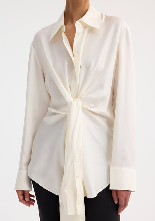 Model wearing Silk knotted shirt | cream, featuring a twisted front design, paired with slim black pants and black strappy heels, showcasing an elegant, refined look.