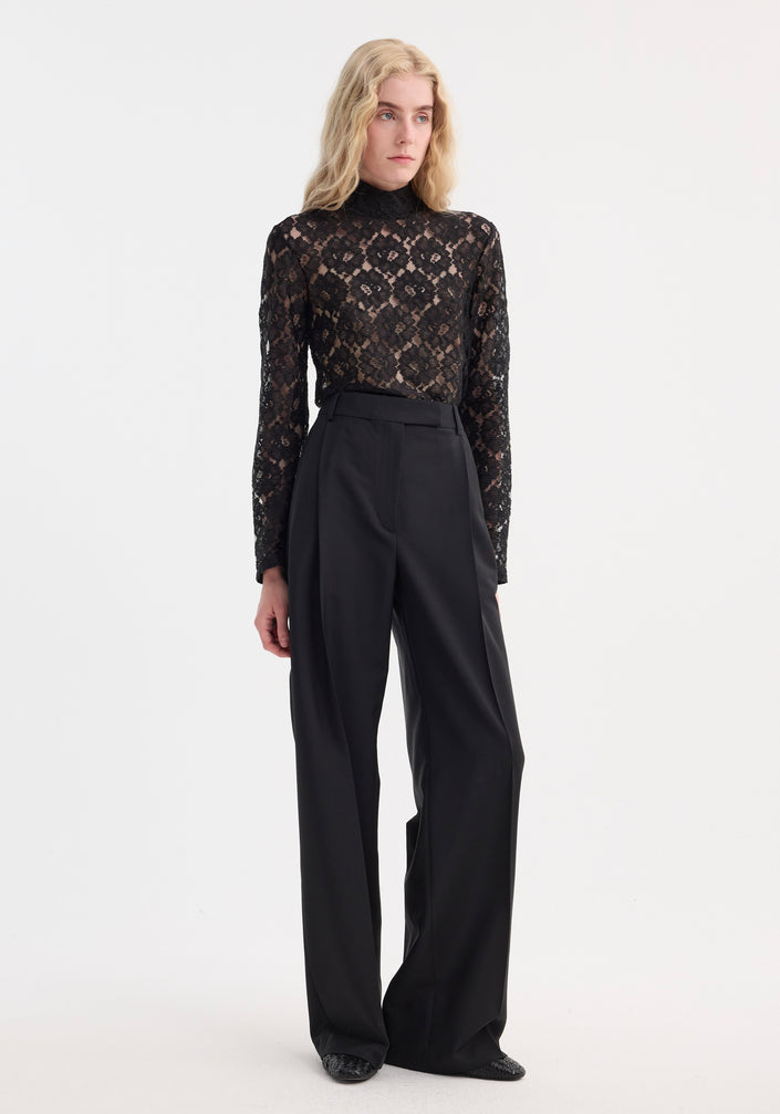 Light-haired person models the Lace turtleneck top | black, featuring a high-neck, long sleeves, and a fitted silhouette, paired with wide-leg trousers.
