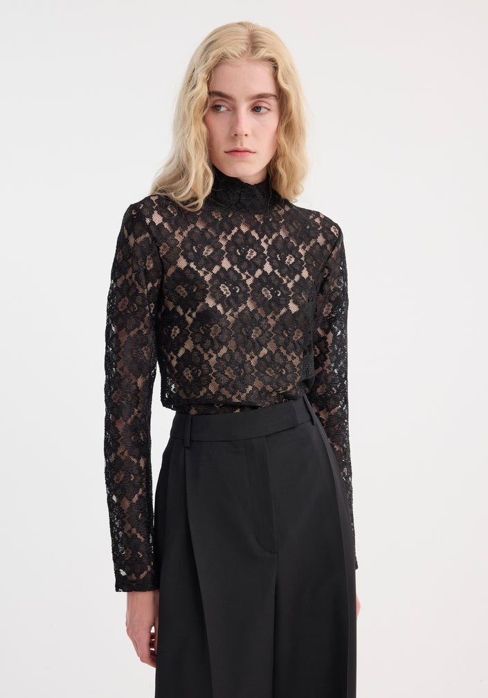 Light-haired person models the Lace turtleneck top | black, featuring a high-neck, long sleeves, and a fitted silhouette, paired with wide-leg trousers.