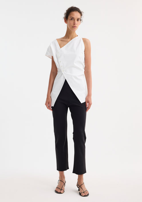 Model in an Asymmetrical poplin top | optic white, featuring an angled hem, off-center buttons, and a side split, paired with black trousers and heeled sandals.