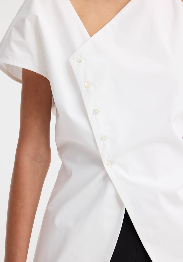 Model in an Asymmetrical poplin top | optic white, featuring an angled hem, off-center buttons, and a side split, paired with black trousers and heeled sandals.