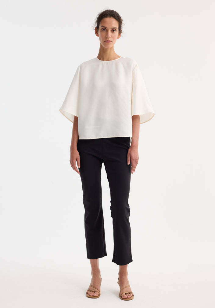 Woman wearing a Buttoned silk top | cream, showcasing a loose fit with elbow-length bell sleeves and back button detailing, paired with slim black trousers.