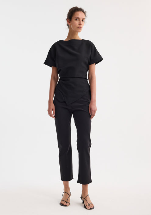 Woman wearing Draped lapel top | black with asymmetrical hem and sculpted shoulders, paired with slim-fitting trousers and strappy sandals, posing fashionably.