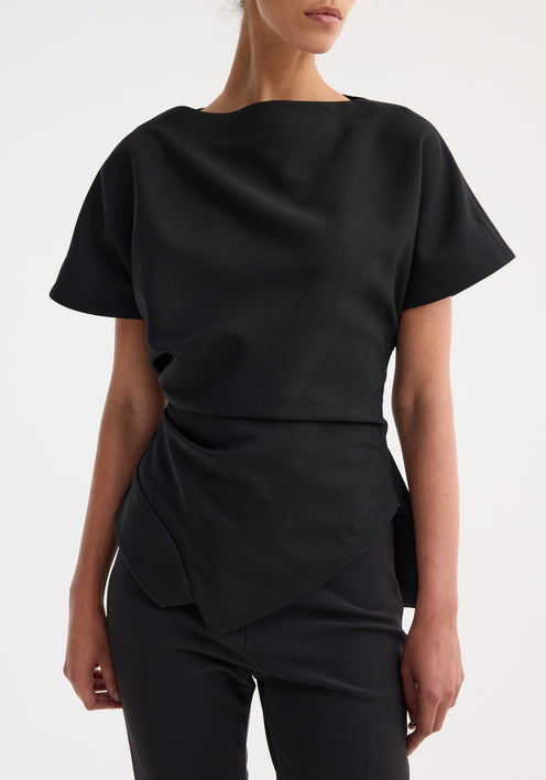 Woman wearing Draped lapel top | black with asymmetrical hem and sculpted shoulders, paired with slim-fitting trousers and strappy sandals, posing fashionably.