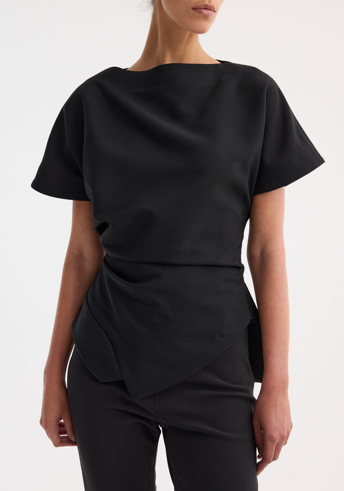 Woman wearing Draped lapel top | black with asymmetrical hem and sculpted shoulders, paired with slim-fitting trousers and strappy sandals, posing fashionably.