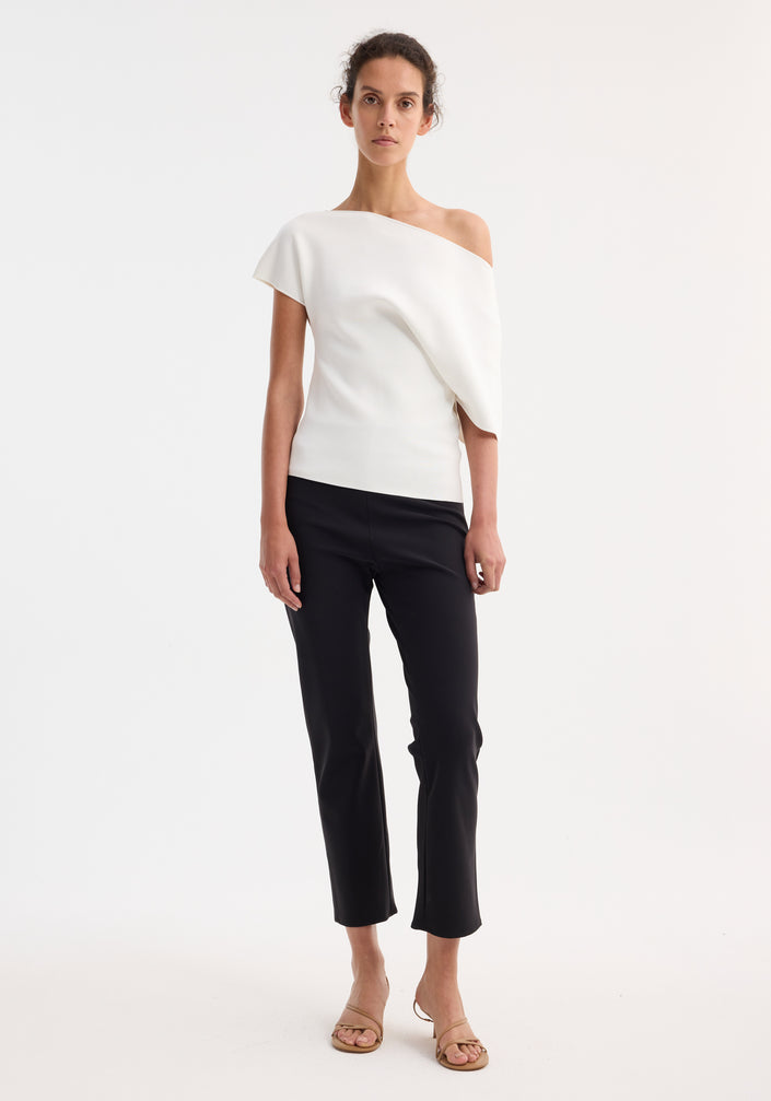 Model wearing Asymmetrical Punto Milano knitted top with slashed neckline and bell sleeve, paired with slim-fit black trousers and tan sandals, hair in updo.