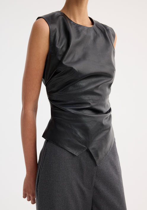 Woman in Sleeveless draped leather top, black, with asymmetrical design and fitted waist, paired with wide-leg trousers and pointed-toe shoes, against white background.