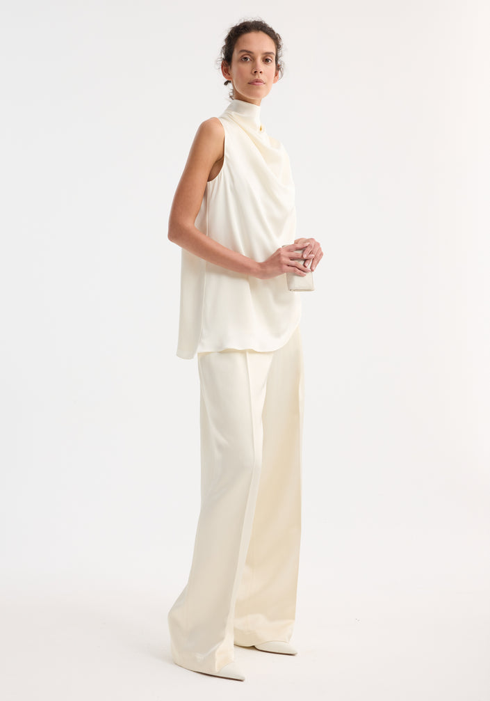 Model wearing Sleeveless draped satin top with cowl neckline, paired with high-waisted trousers and holding a clutch, showcasing minimalist, elegant style.