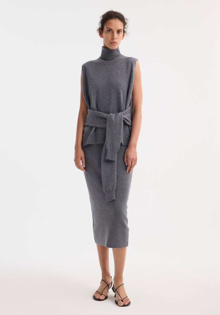 Model wears Double-layered knit dress in mid grey melange, featuring a sleeveless turtleneck top, matching midi skirt, and sweater tied around waist, with strappy sandals.