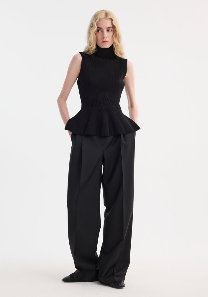 Model wears Sculptural sleeveless knitted top with peplum flare, crafted from wool-cashmere. Paired with high-waisted, wide-leg trousers, hands in pockets.