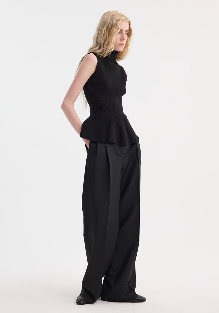 Model wears Sculptural sleeveless knitted top with peplum flare, crafted from wool-cashmere. Paired with high-waisted, wide-leg trousers, hands in pockets.
