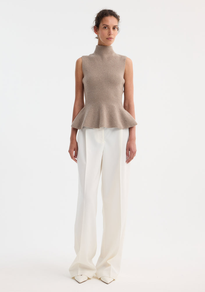 Model wearing the Sculptural sleeveless knitted top in taupe melange, featuring a high neckline and peplum flare, paired with high-waisted, wide-leg trousers.
