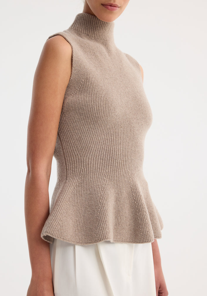 Model wearing the Sculptural sleeveless knitted top in taupe melange, featuring a high neckline and peplum flare, paired with high-waisted, wide-leg trousers.