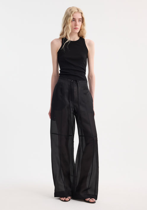 Model in Organza silk parachute trousers, showcasing wide-leg, semi-sheer flow and drawstring waist, paired with black tank top and strappy sandals.