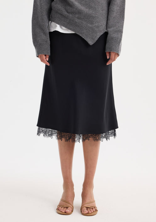 Woman wearing Lace-trimmed silk skirt | black, featuring delicate lace at hem, paired with a heather-gray V-neck sweater.