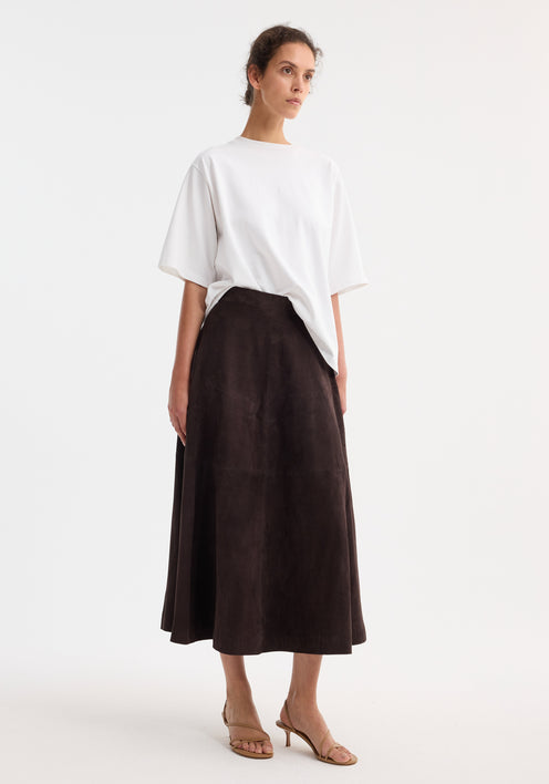 A-line suede skirt in dark chocolate, featuring a soft, voluminous shape. Model pairs it with a white T-shirt and tan sandals for a minimalist look.