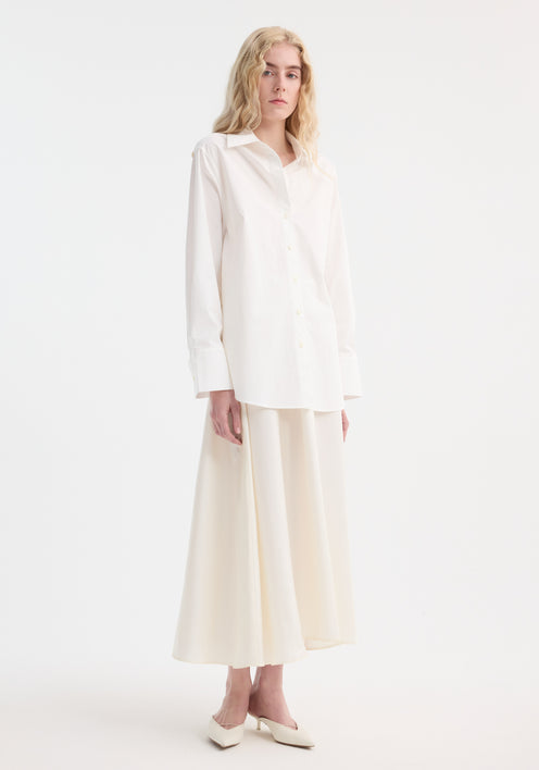 Young adult models the Poplin silk combined gilet-dress, featuring open armholes and a buttoned wrap belt, creating a fluid silhouette with cotton and silk elements.