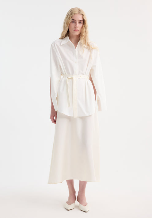 Young adult models the Poplin silk combined gilet-dress, featuring open armholes and a buttoned wrap belt, creating a fluid silhouette with cotton and silk elements.