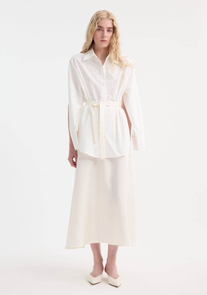 Young adult models the Poplin silk combined gilet-dress, featuring open armholes and a buttoned wrap belt, creating a fluid silhouette with cotton and silk elements.